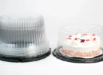 "A frosted cake with colorful sprinkles and strawberry topping in a JC Packaging clear cake container, beside an empty stack of clear containers, all against a clean white background."