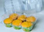 "JC Packaging's Six-Muffin Display Case, with half-open lid revealing six scrumptious yellow muffins in bright festive liners, set against a backdrop of stacked cases."